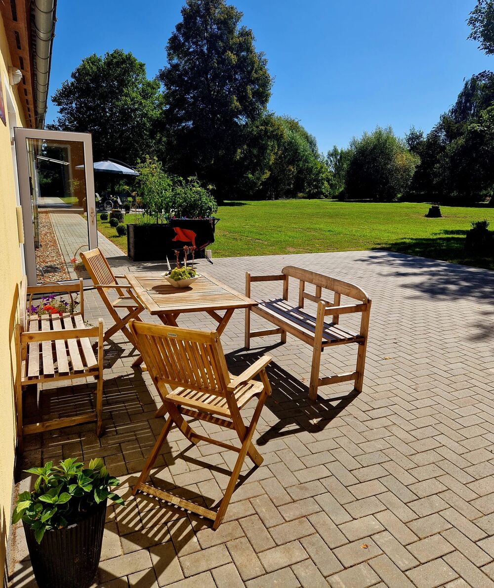 Sonnige Terrasse mit Holzmöbeln, dahinter die weite Rasenfläche des Gemeinschaftsgartens