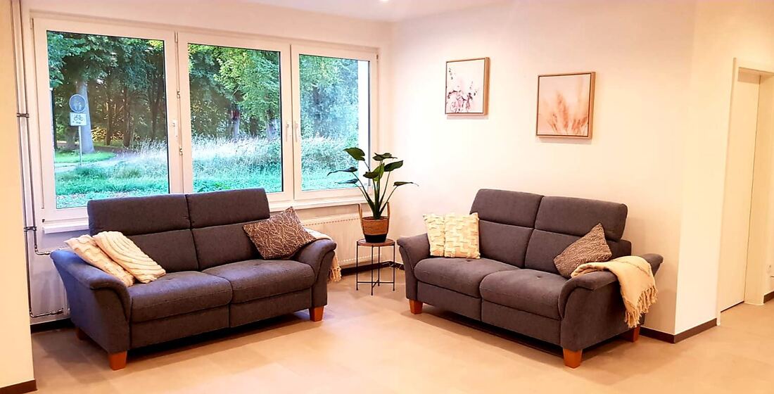 Gemeinschaftliches Wohnzimmer mit zwei Sofas, Decken und Blick durch große Fenster ins Grüne