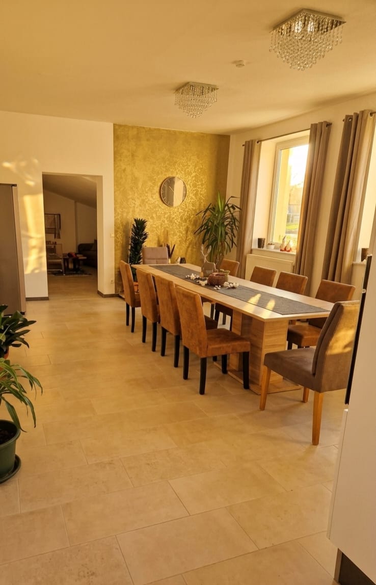 Gemeinschaftsraum im betreuten Wohnen: lange Tafel mit gepolsterten Stühlen vor einer goldenen Tapetenwand, darüber Kristall-Deckenleuchten